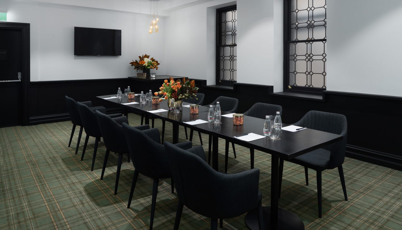 Long dining tables with black chairs, place settings, and flowers in a room with tall windows and patterned carpet.