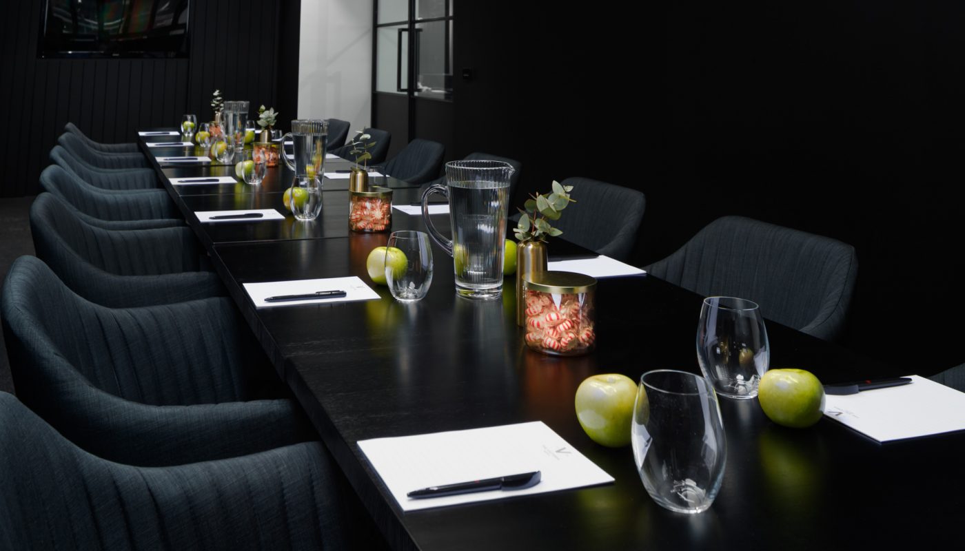 A long black conference table lined with chairs, glasses, fruit, snacks, and notepads in a dim room.