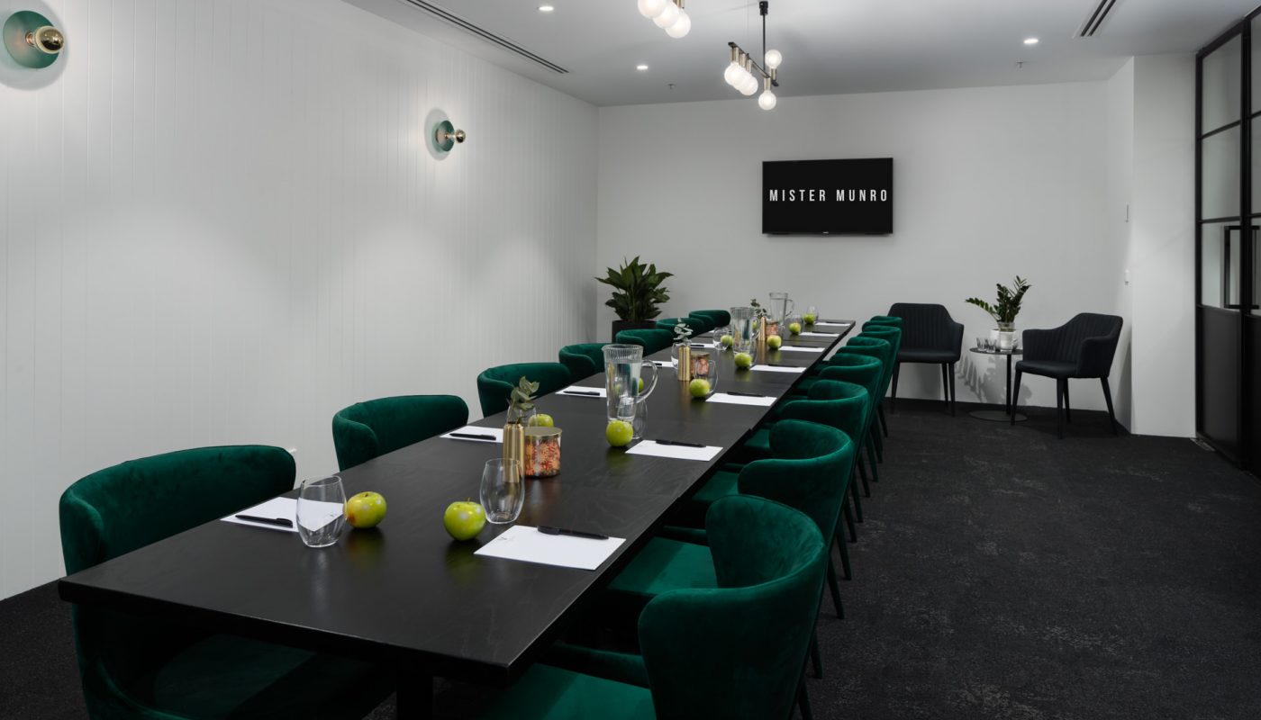 Modern meeting room with a long black conference table, green upholstered chairs, water bottles, and small plants.