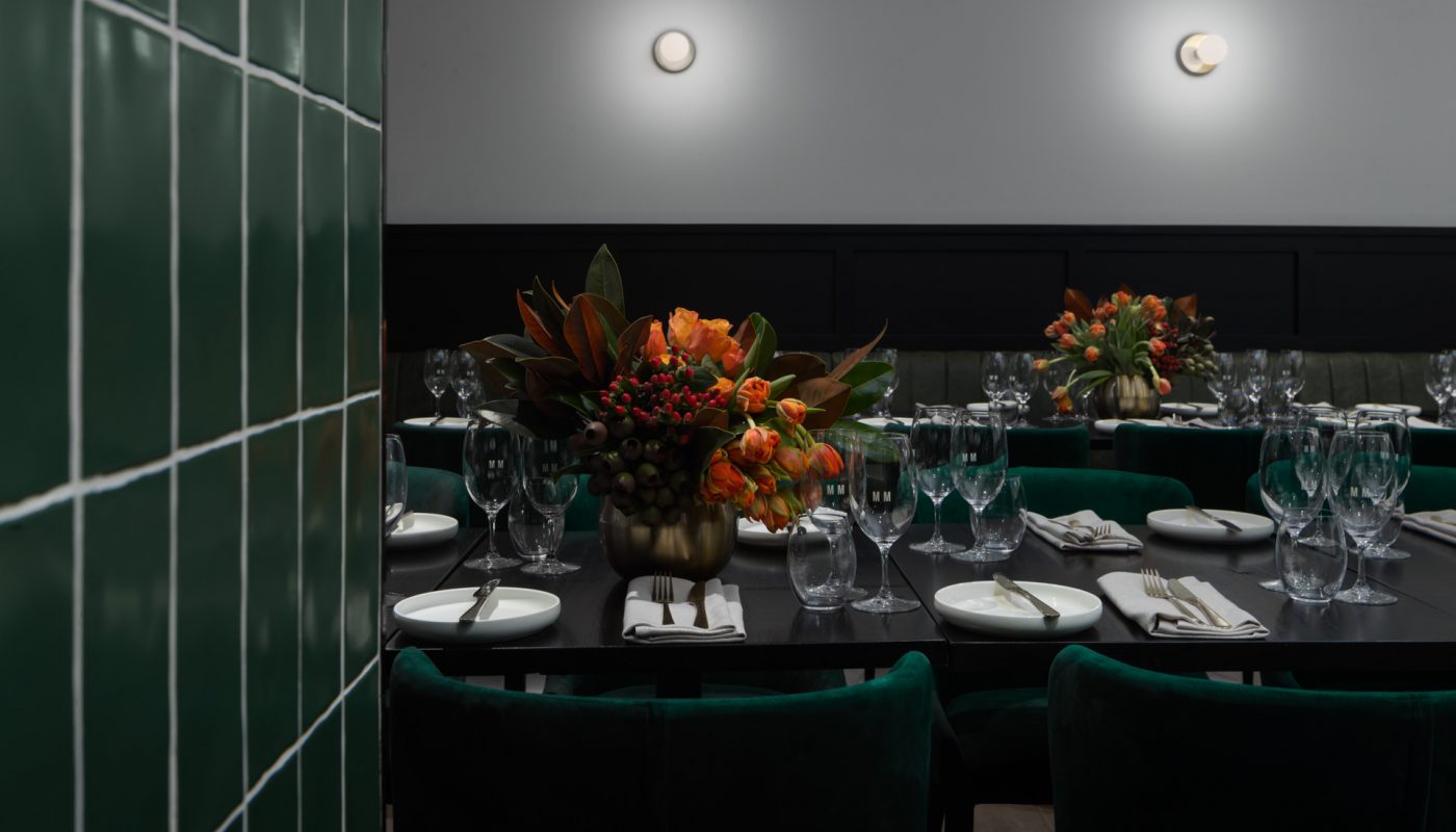 A dim dining room with set tables, glassware, floral arrangements, and a dark green tiled wall.