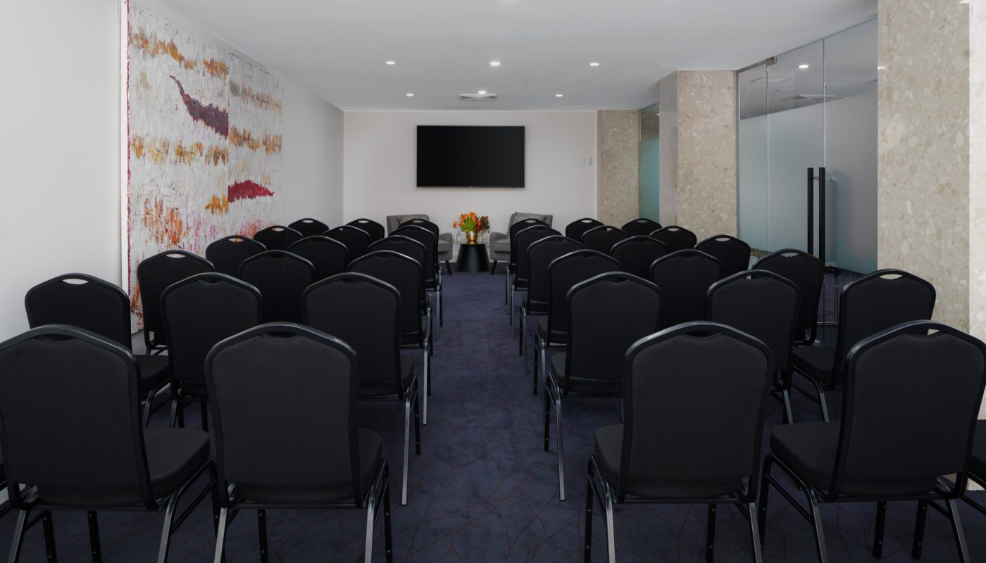 Rows of black chairs line a conference room with a central aisle facing a wall-mounted screen.