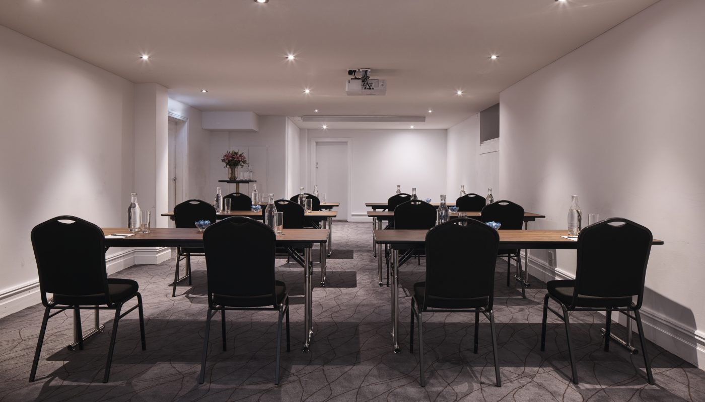 An empty conference room with rows of tables and chairs, recessed ceiling lights, and a ceiling-mounted projector.